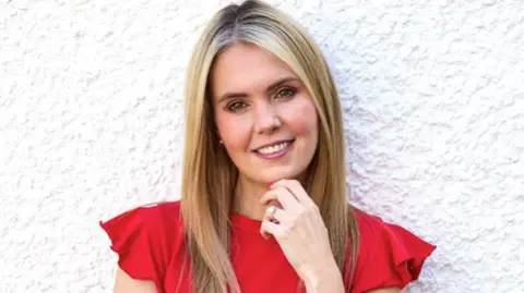 Canseecantsee Claire Sisk, standing in front of a white wall, she is wearing a red top, she has her hand to on her chin, with a ring on the second finger. She has long, blond hair, straight hair, is smiling straight to the camera with her head slightly to the left.
