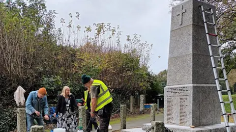 Terri Beer Four people on the left looking to the ground with cleaning equipment. There are two people wearing hi-vis on to the left, a woman at the centre wearing a dress and a man in a denim jacket. On the left is the grey war memorial with names at the bottom. There is a ladder against it on the right.