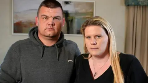 Lee and Micala Trussler stand side-by-side in a room with a painting on the wall behind them. Lee has short dark hair and is wearing a grey hoodie, Micala has long blonde hair and is in a black top.
