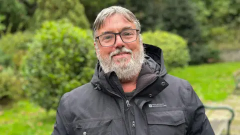 A man wearing a black coat, looking towards the camera wearing black glasses. He has grey hair with a middle parting and a grey beard.