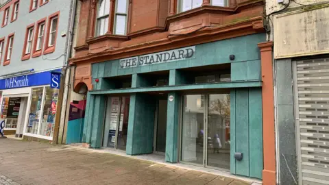BBC A green-fronted town centre property with sandstone surrounds sits between one empty building and a WH Smith store