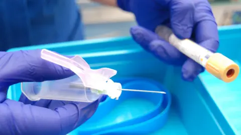 Kettering General Hospital A blue gloved- hand holding a syringe with a needle, and another gloved hand holding a sample tube. They are being held over a blue tray.