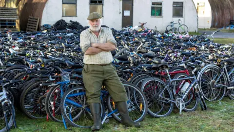 Graeme Hart / PPA Nigel Carter leans against over a thousand bikes with his arms crossed looking at the camera. He has white hair and a beard, wearing a checked shirt and green trousers with black welly boots.