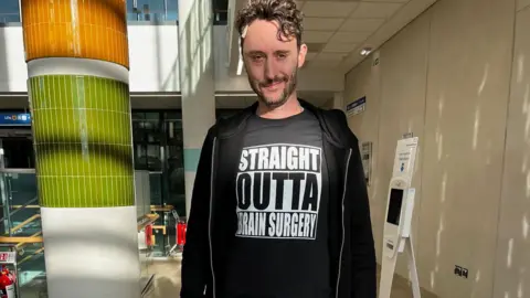 Matthew White Matthew White in hospital, wearing a hoody, PJ bottoms and a t-shirt saying STRAIGHT OUTTA BRAIN SURGERY.