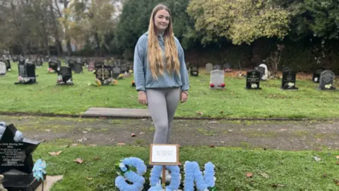 A blonde woman is wearing a grey hoody jumper and grey leggings. She is standing behind a blue wreath that says son, in a blue floral arrangement