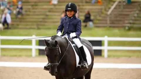 Kavitha Govind in a navy suit jacket, white trousers, black boots and black riding hat. He sits on top of a dark brown horse in a riding competition ring.