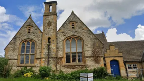 Google An old schoolhouse, built in Cotswold stone, with two pointed gables, large windows and a central chimney-like pillar between the two. There is a door painted blue besides one of the gables. There is a green space with grass and bushes in front of the building, and a sign. It is a partly cloudy day, with sections of blue sky and cloud.