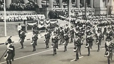 Handout A black and white photo showing Singaporean girls in a marching band performing at an event. Officials, and the Singapore flag, can be seen in the background