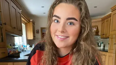Family handout Jeni Larmour smiling in a kitchen. She's wearing a red and black sweatshirt and has light brown curly hair with dark eyebrows.