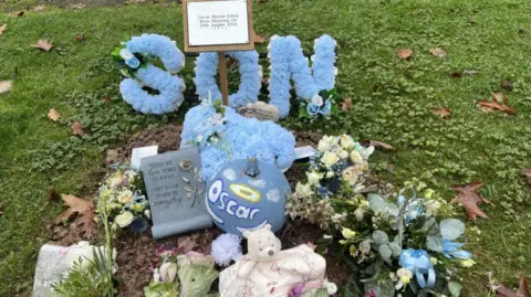 BBC A small grave, is surrounded by grass. There is a blue wreath that says son in blue flowers and a blue pumpkin that says Oscar with a yellow halo above the name.