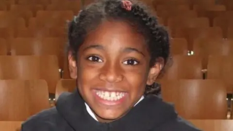 Family handout Ella Adoo-Kissi-Debrah with big smile, wearing a black jumper with rows of chairs in the background