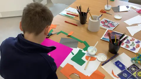 Anna Mears A child sitting with it's back to the camera so you can not see their face. They are sitting in a classroom at a school desk cutting out paper shapes on coloured cards.
