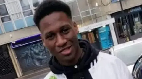 Avon and Somerset Police A young man with black afro hair. He is wearing a black hoodie with a white jacket. Behind him is a load of shops.