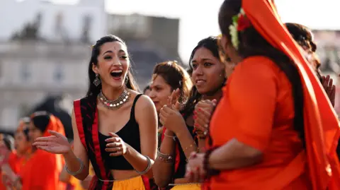 PA Media Women performers in black and orange clothing clap and chat in Trafalgar Square