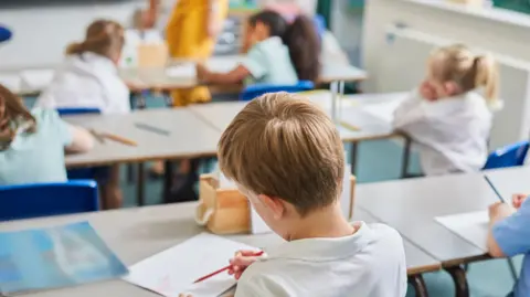 Getty Images A group of children in a classroom sitting on blue chairs with their backs to the camera. They are wearing either white shirts or green dresses and have paper and pencils on their desks.