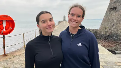 Lois Vidamour and Jenny Thomas smiling at the St Peter Port lighthouse. They are dressed in running quarter zips and their hair is tied back.
