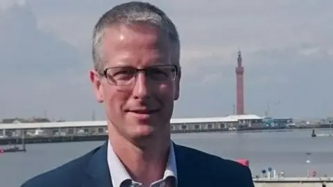 North East Lincolnshire Council Rob Walsh with short-cropped grey hair. He is wearing dark-metal framed specs on his face, a navy blazer over a white and blue thin-striped shirt. He is standing on the dock with Grimsby's Dock Tower in the background and a row of one-storey buildings.
