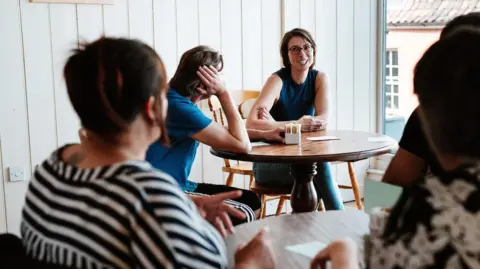 Rob Dodsworth A group of people at a coaching session, in a room with a table, and talking to each other