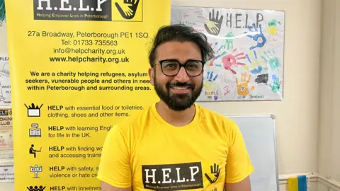 Smiling at the camera is Bilal Aslam, who is wearing a black circle rimmed glasses and a bright yellow t-shirt with the words H.E.L.P. printed on a black text box. He has black hair, a black beard and is posing for the photo
inside an office. Behind him is an advert for the charity.