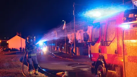 PA Media A firefighter dampens down his own truck due to extreme temperatures as a huge bonfire in Craigyhill, Larne, is lit on the "Eleventh night" to usher in the Twelfth commemorations.