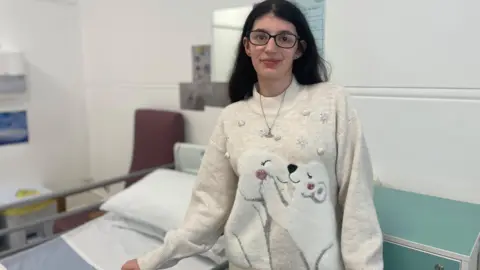 SimonThake A young girl in a white jumper stands infront of a hospital bed
