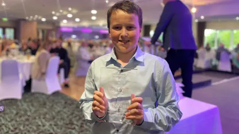 Hamish holding his carer award at the ceremony. He is wearing a pale-blue shirt and smiling. There are people in the background sitting at tables.