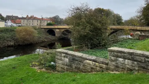 BBC A bridge over a river with grass banks on either side and a stepped brick wall going up the right hand side of the bank