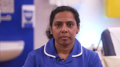 Looking directly at the camera is Tincy Jose wearing her blue nursing uniform which has a white lining around the collar. She has dark hair, brown eyes and white stud earrings. She is inside the Queen Elizabeth Hospital in King's Lynn and behind her there is a hospital bed and sink.