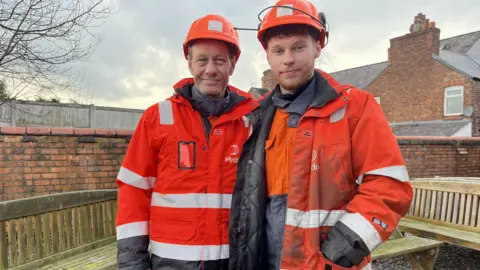 BBC Two men dressed in orange hi-vis clothing with a logo that reads Hydro and wearing orange hard hats stand in a walled outsdie space in front of some oak benches. One of them is a man in his 50s and the other, a younger man of 21 with a short ginger beard.