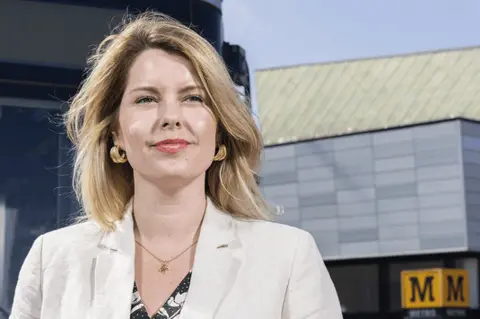 North East Combined Authority (NECA) The North East Mayor Kim McGuinness. She has shoulder length blonde hair and is wearing large gold earrings and a cream coloured suit. She is standing outside a Tyne and Wear Metro station.
