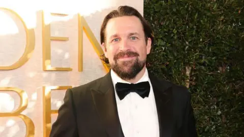 Getty Images Trevor Matthews is looking at the camera, he is stood in front of a sign which says Golden Globes in gold font, but you can only read the 'D E N' of golden and the 'B E' of globes. He is wearing a black tuxedo with a black bow tie and a white shirt. He is smiling at the camera and his dark hair has a middle parting, he has a moustache and beard in the style of a goatee.