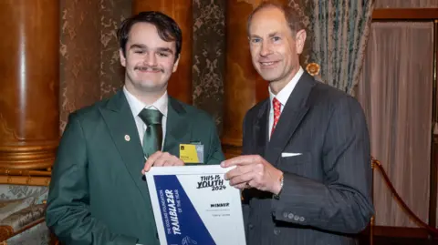 DofE/Jonny Sanders Harry Jones (elft) being handed an award by the Duke of Edinburgh, Harry is wearing a green suit, with a green tie and white shirt, he has short dark hair and a moustache, the Duke is wearing a grey pinstriped suit with a red tie, they are standing by pillars and curtains. Both men are smiling and looking straight at the camera.
