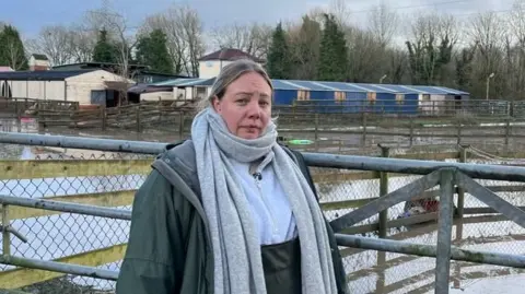 Davinia Ramos/BBC A woman wearing overalls, a jacket and a big scarf stands in front of a flooded field