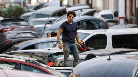 EPA A man wearing mud-stained clothing stands on a pile of cars swept down a road by flood waters in Valencia, in Spain in November 2024