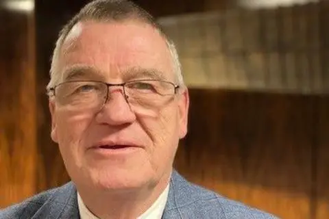 The Leader of Gateshead council, Martin Gannon, pictured inside Gateshead civic centre. He has short grey hair and is wearing thin-rimmed glasses. He is wearing a grey checked suit.