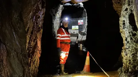 A man in hi-visability clothing stands in a mine shaft. He is wearing a hard hat with a head torch. He is looking back towards the camera, framed by rock walls, with the shaft continuing on behind him up to a gate that has fenced it off.