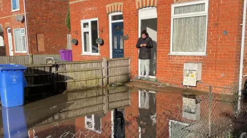 A flooded garden with a woman standing in a doorway cut off by water taking pictures of the garden