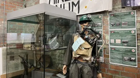 A mannequin wearing the kind of kit which would have been worn by the mine rescuers, khaki clothing and a dark green hard hat and breathing apparatus. There is a display cabinet to its left with some mining memorabilia and to its right, a brick wall with information boards about pits in north Wales.