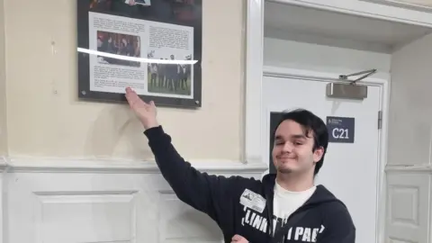 Harry Jones Harry pointing at a picture of his Duke of Edinburgh award in a hallway, he is smiling and looking at the camera, wearing a black hoodie, and white top. He is by a white door, and walls with panelling on.