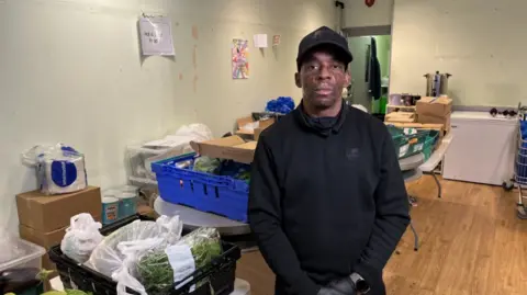 BBC Dean Foster, a young men wearing a back cap and black clothes, standing in a room in front of boxes of fresh food