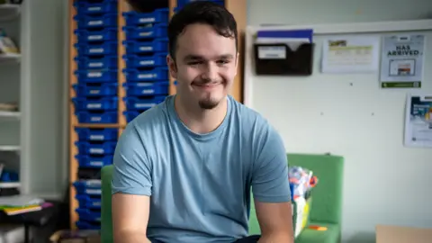 DofE/Jonny Sanders Harry smiling and looking straight at the camera, wearing a blue T-shirt, in a classroom with shelves behind him. He has short dark hair and a goatee beard.