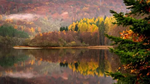 PA Media A photograph of a loch during autumn. Trees with red, orange and yellow leaves cover a hillside and reflect onto the calm water.