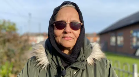 BBC Dureshahwar Shaikh is wearing a green coat with a furry hood. She has sunglasses covering her eyes and a black hijab on her head.