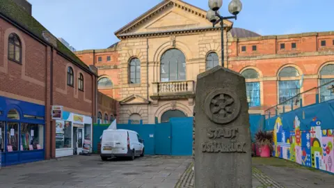 BBC A stone monument in front of Derby's market hall. Behind the stone and in front of the market hall is blue temporary fencing.